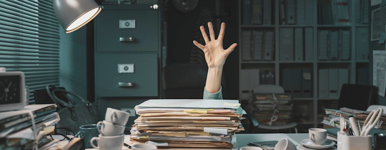 Ein voller Schreibtisch steht in einem Büro. In der Mitte des Bildes ist eine ausgestreckte Hand. Es ist ein Symbolbild für jemanden, der in Arbeit ertrinkt.