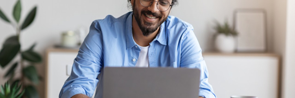 Junger Mann mit dunklen, längeren Haaren und hellblauen Hemd sitzt vor Laptop.