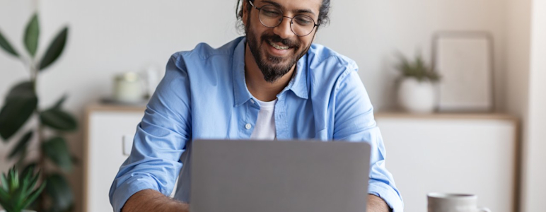 Junger Mann mit dunklen, längeren Haaren und hellblauen Hemd sitzt vor Laptop.