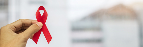 Eine Hand hält eine rote Schleiße, das Symbol für Solidarität mit HIV-Infizierten und AIDS-Erkrankten.
