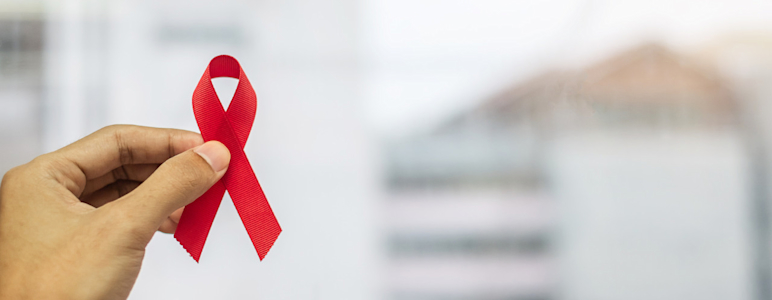 Eine Hand hält eine rote Schleiße, das Symbol für Solidarität mit HIV-Infizierten und AIDS-Erkrankten.