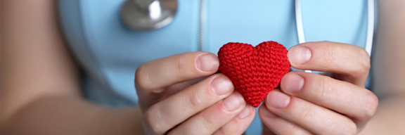 Frau mit Stethoskop hält ein gehäkeltes Herz in den Händen.