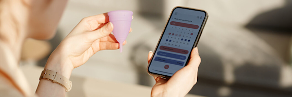 Frau hält Menstruationstasse in der linken Hand und Handy mit Zykluskalender in der rechten.