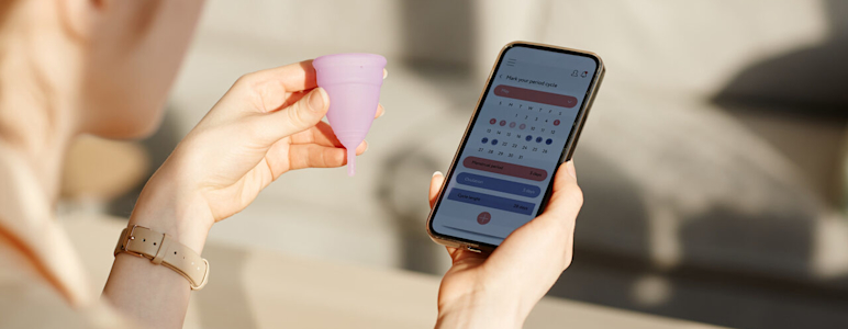 Frau hält Menstruationstasse in der linken Hand und Handy mit Zykluskalender in der rechten.