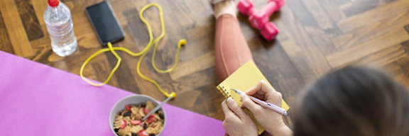 Kombination aus Ernährung und Sport um abzunehmen. Junge Frau schreibt in ein Notizbuch, sitzt auf Yogamatte und hat gesundes Müsli und Hanteln vor sich.