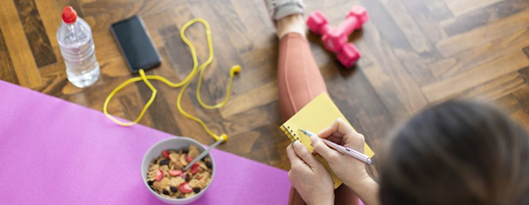 Kombination aus Ernährung und Sport um abzunehmen. Junge Frau schreibt in ein Notizbuch, sitzt auf Yogamatte und hat gesundes Müsli und Hanteln vor sich.