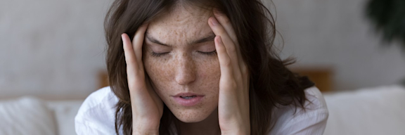 Junge Frau mit Sommersprossen hat Kopfschmerzen und hält sich den Kopf.