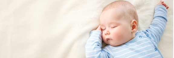 Baby mit blauem Strampler schläft auf heller Decke.