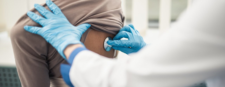 Ein Arzt hört mit dem Stethoskop am Rücken einer Patientin die Lunge ab.