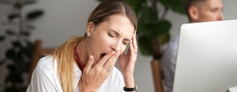 Eine Frau ist müde im Büro und gähnt an ihrem Schreibtisch am Laptop.