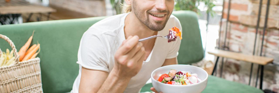 Auf dem Bild ist ein junger Mann zu sehen, der auf einem grünem Sofa sitzt und einen Salat isst.
