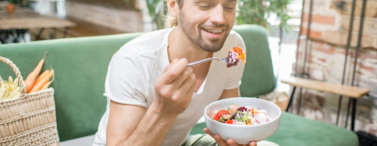 Auf dem Bild ist ein junger Mann zu sehen, der auf einem grünem Sofa sitzt und einen Salat isst.