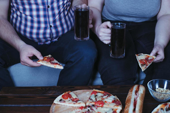 Übergewichtiger Mann und übergewichtige Frau sitzen auf dem Sofa und essen Pizza und trinken Cola.