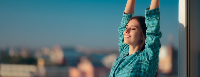Junge Frau mit kariertem Hemd streckt sich in der Sonne. Im Hintergrund ist die Skyline einer Stadt zu sehen.