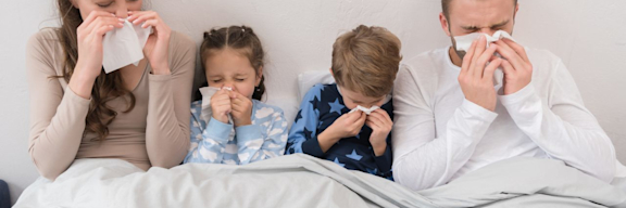 Familie ist zusammen im Bett. Alle sind krank und schnäuzen in ein Taschentuch.