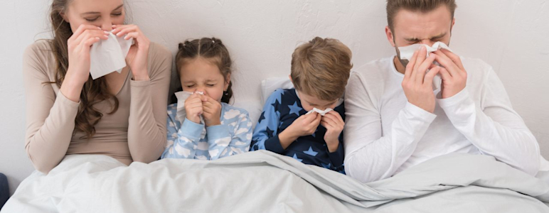 Familie ist zusammen im Bett. Alle sind krank und schnäuzen in ein Taschentuch.