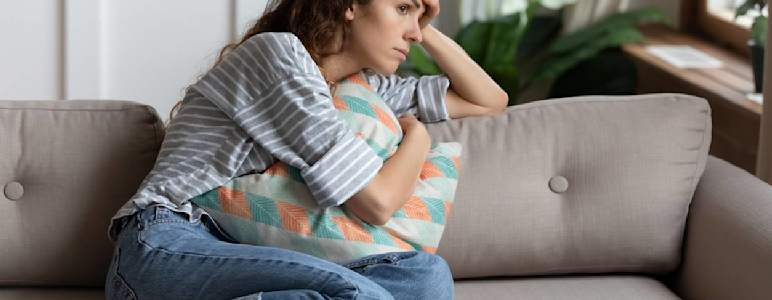 Frau sitzt auf dem Sofa und hält sich die Stirn mit einer Hand und ein Kissen mit der anderen Hand