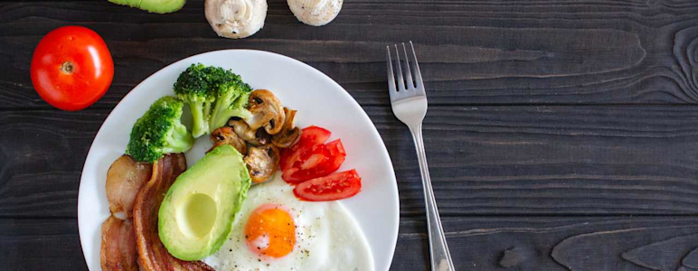 Es ist ein Teller mit ketogenen Lebensmitteln zu sehen, wie Avocado, Tomate, Pilzen, Broccoli, Speck und Ei.