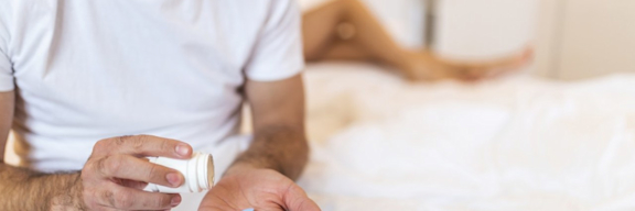 Mann sitzt auf Bett mit Viagra Pille auf der Hand.
