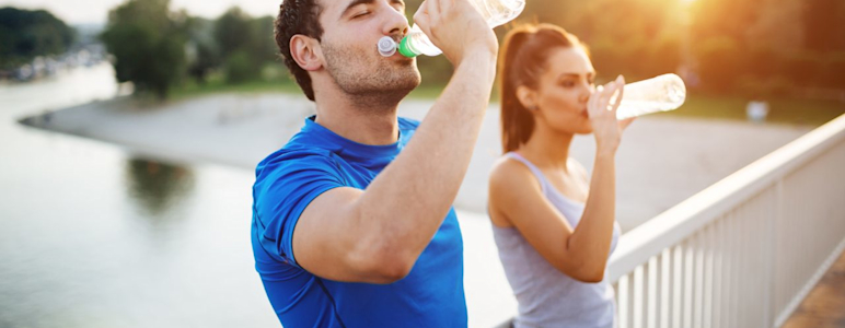 Es ist ein junges Paar in Sport-Outfits zu sehen, beide trinken Wasser.