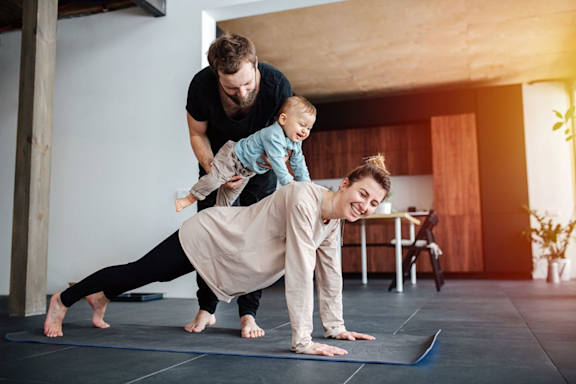 Sportliche Aktivität zu Hause mit der Familie. Frau macht Workout auf Yoga-Matte, Mann und Baby sind dabei.