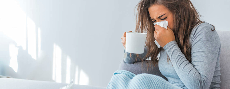 Eine erkältete Frau hält eine Teetasse in der einen Hand und putzt sich mit der andere Hand die Nase.