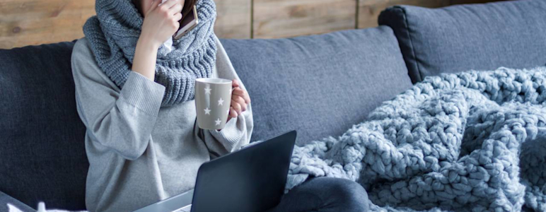Junge Frau sitzt auf der Couch und putzt sich die Nase. Auf ihrem Schoß steht ein Laptop.