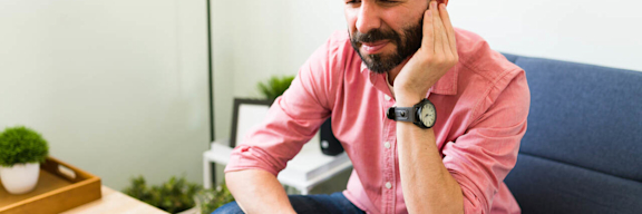 Mann sitzt auf einem Sofa und hält sich das Ohr mit schmerzverzerrtem Gesicht.