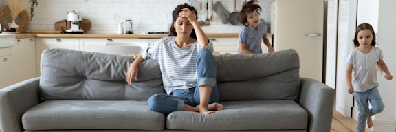 Eine Frau sitzt genervt auf dem Sofa, während Kinder um sie herumtoben.