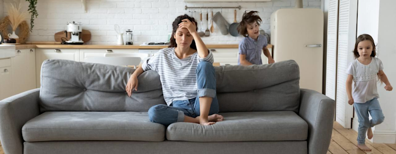 Eine Frau sitzt genervt auf dem Sofa, während Kinder um sie herumtoben.