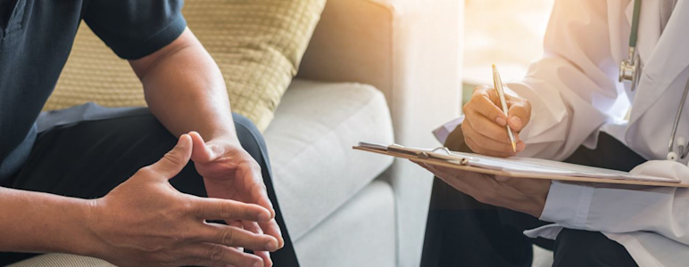Ein Patient sitzt auf der Couch, ihm gegenüber sitzt ein Arzt, der sich Notizen macht.