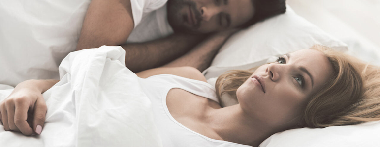 EIne junge Frau liegt wach im Bett und starrt nach oben, daneben schläft ein Mann.