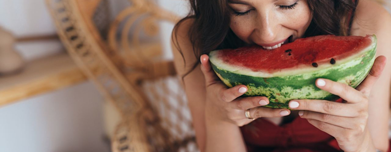Es ist eine junge Frau mit dunklen Haaren zu sehen, die einen Strohhut trägt und in ein großes Stück Melone beist.