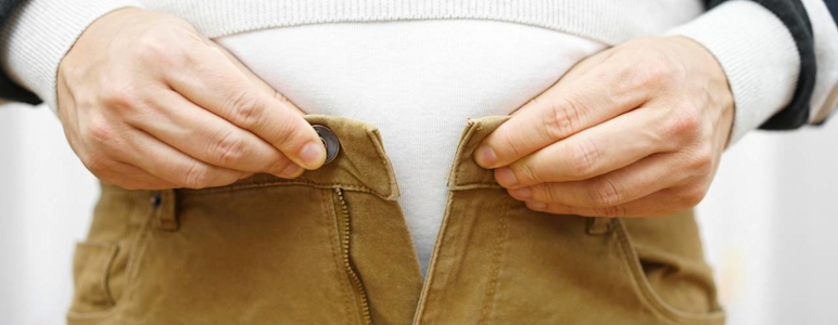 Person bekommt Knopf an der braunen Hose über dem Bauch nicht geschlossen.