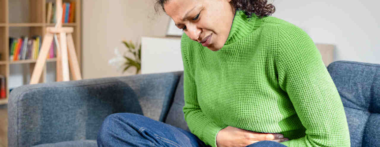 Frau sitzt zusammengekrümmt auf dem Sofa und hält sich den schmerzenden Bauch.