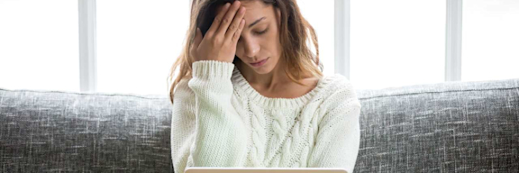 Eine Frau sitzt mit Laptop auf dem Sofa und hat Kopfschmerzen.