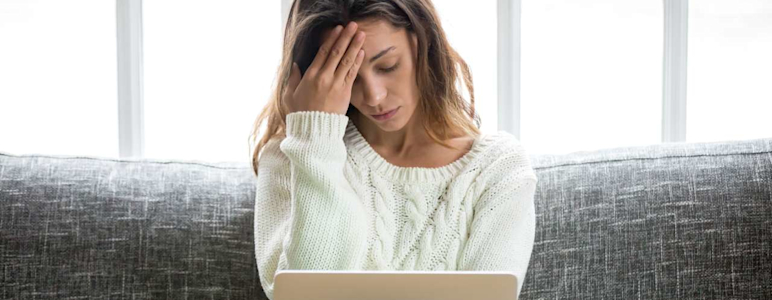 Eine Frau sitzt mit Laptop auf dem Sofa und hat Kopfschmerzen.