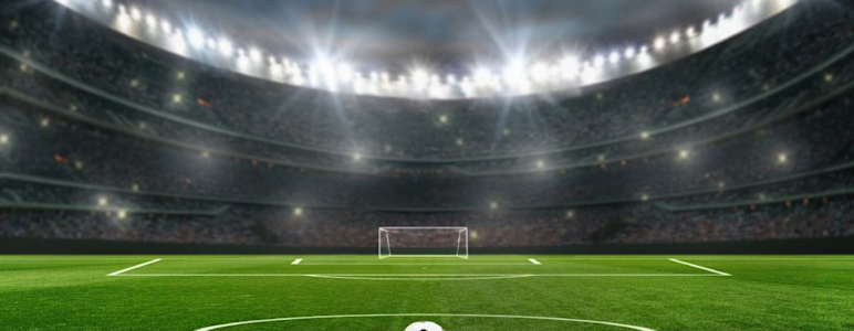 Fussball liegt im Stadion vor der Mittellinie. Im Hintergrund Flutlicht und ein leeres Tor.