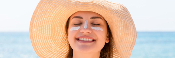 Junge Frau am Meer, die einen Sonnenhut trägt und Sonnencreme im Gesicht hat.