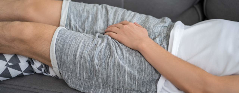 Ein Mann liegt auf dem Sofa und hält sich den schmerzenden Genitalbereich.