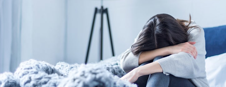 Junge Frau in grauer Kleidung sitzt zusammengerollt auf dem Bett zu Hause.