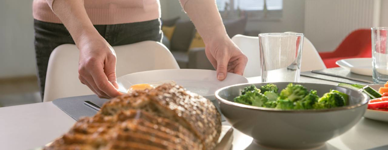 Im Bild ist ein gedeckter Tisch zu sehen, darauf aufgeschnittenes Brot und Broccoli. Eine Frau stellt einen Teller auf den Tisch.