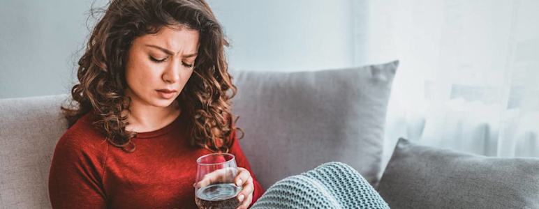 Eine Frau sitzt auf dem Sofa und sieht beunruhigt aus. Sie hält ein Glas Wasser und eine Tablette in den Händen.