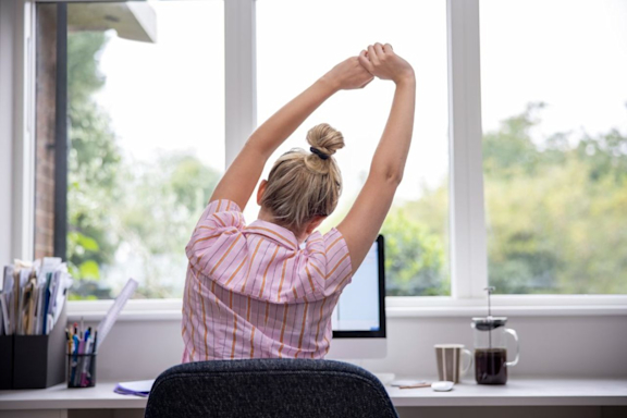 Frau im Büro oder Homeoffice hat Rückenschmerzen und streckt ihre Arme.