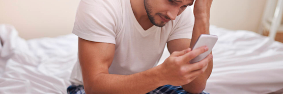 Mann sitzt auf Bettkante und schaut traurig und verzweifelt auf sein Handy.