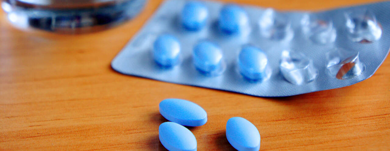 Blaue Tabletten in einem Blister liegen auf einem Holztisch, im Hintergrund steht ein Glas Wasser.