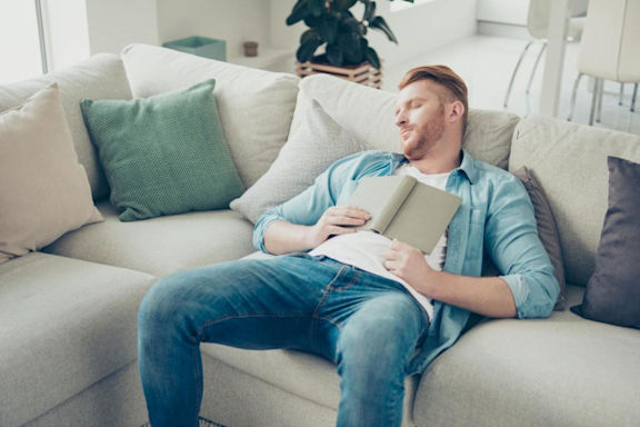 Mann mit Boreout ist gelangweilt und schläft mit Buch auf Sofa.