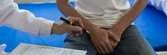 Ein Mann sitzt beim Arzt auf der Liege und hält sich vor Schmerzen den Intimbereich.