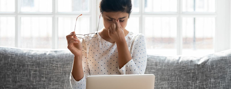 Eine junge Frau sitzt auf der Couch, vor ihr steht ein Laptop. Sie hat ihre Brille abgenommen, schließt die Augen und wirkt gestresst.