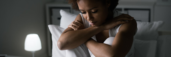 Eine junge Frau sitzt niedergeschlagen auf einem Bett.
